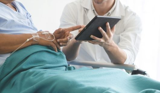 A patient sits up in a hospital bed and points at a tablet being held by a clinician sitting beside them.